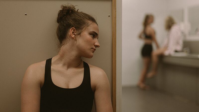 A teenage girl with hair in a bun sits on the ground, while two out-of-focus can be seen talking to each other.