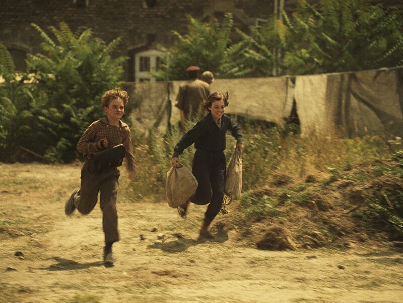 Two children run along a grassy road.