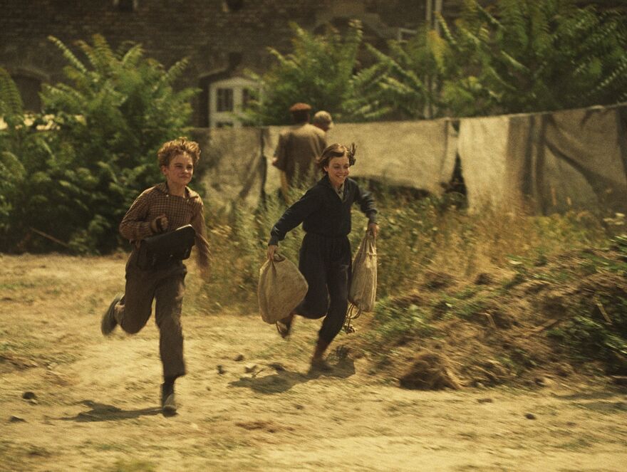 Two children run along a grassy road.