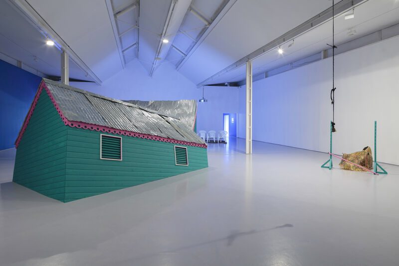 An installation image of gallery two showing a green structure like a building with a corrugated roof and a pink trim, as though it is sinking into the floor.