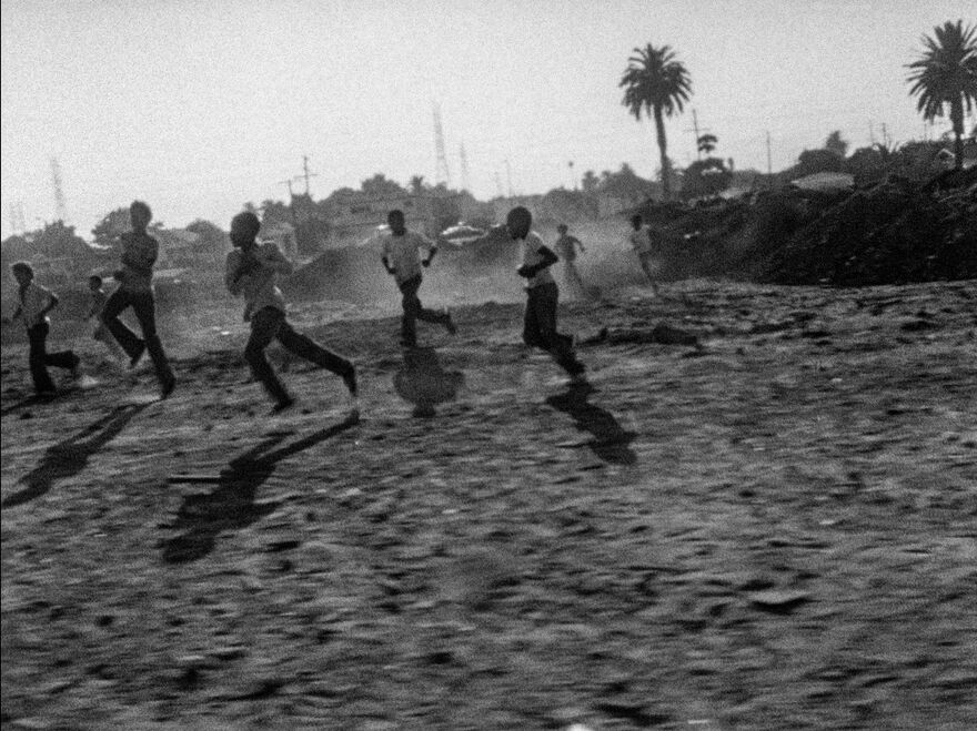 A slightly out of focus black and white image of young boys playing on a sandy beach palm trees visible in the background