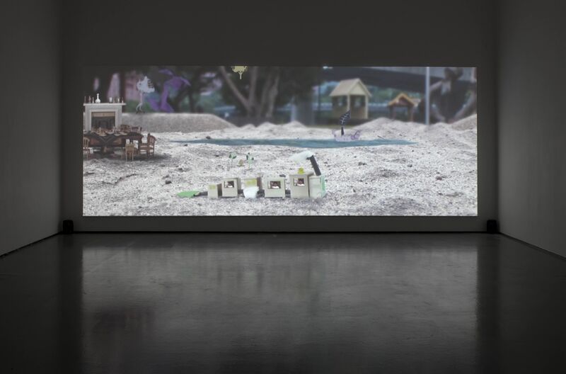From Hiraki Sawa's exhibition. A screen shows an image of a miniature train made out of cardboard in a sand-pit. A miniature dining table with chairs and a fireplace also sits in the background of the sandpit.