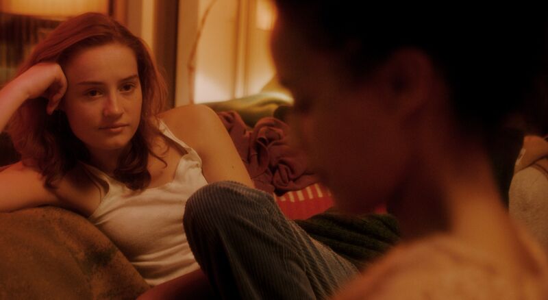 A teenage girl sits on a couch looking at another woman who is blurred.