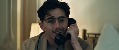 A young man wearing glasses smiles while talking on an old-fashioned phone