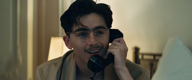 A young man wearing glasses smiles while talking on an old-fashioned phone
