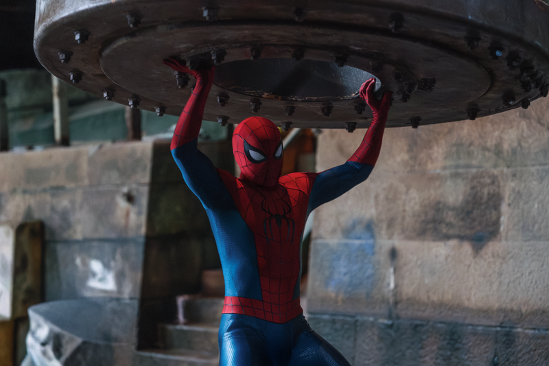 Spider-Man holds a large metal disk above his head while standing in front of a stone wall with stairs.