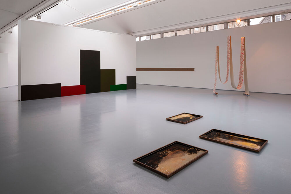 A gallery space with wallpaper suspended from the ceiling and a mural on the wall, with framed pictures on the floor 