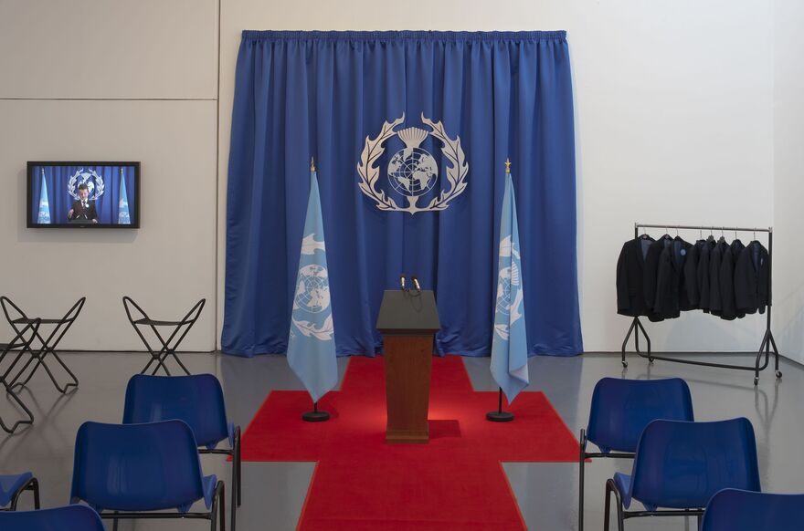 From 'Imagine Being a World Leader'. A podium for making speeches is in the middle of DCA Galleries. There are two flags with a logo that looks like the UN logo on it. There is a TV in the background with a child making a speech, and some small black blazers to borrow.