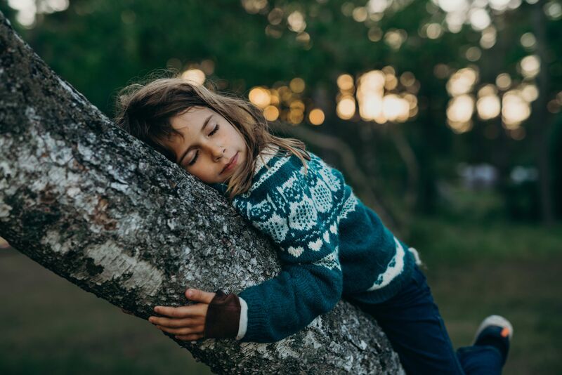 A young child in a jumper hugs a tree with her eyes closed.
