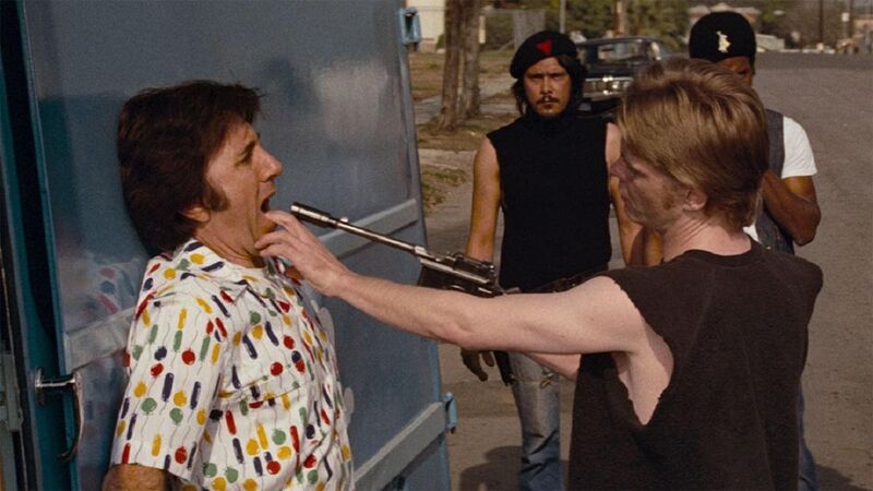 Man holding a gun to another man's face as he stands with his back against a van. Two other men look on