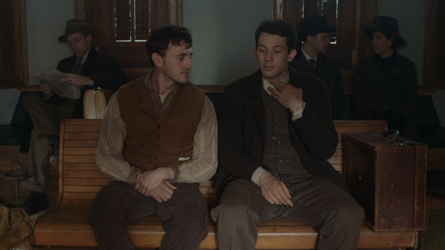Two men in 1940s clothing sit together on a bench in a train station