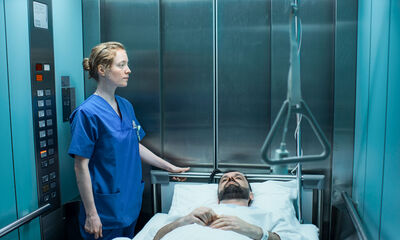 A nurse in a blue uniform stands in a lift with a patient lying on a bed.
