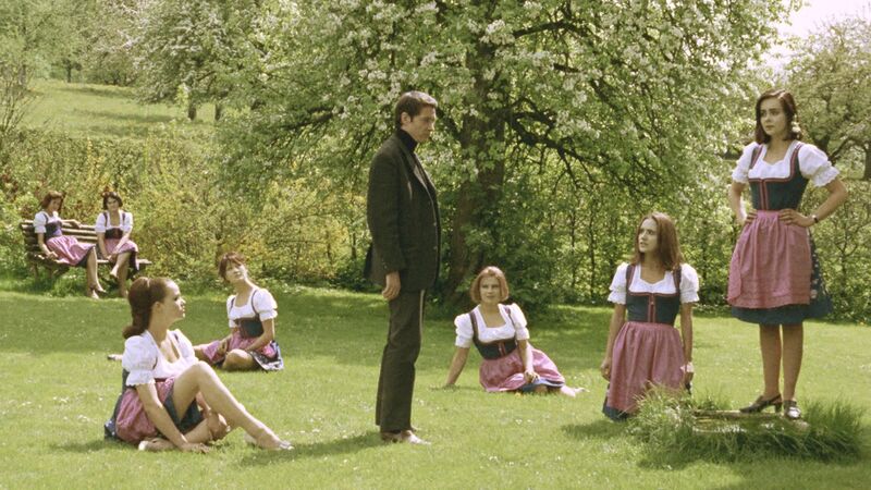 A group of women wearing dirndls stand and sit in a green field, with one man in a suit standing in the middle of them