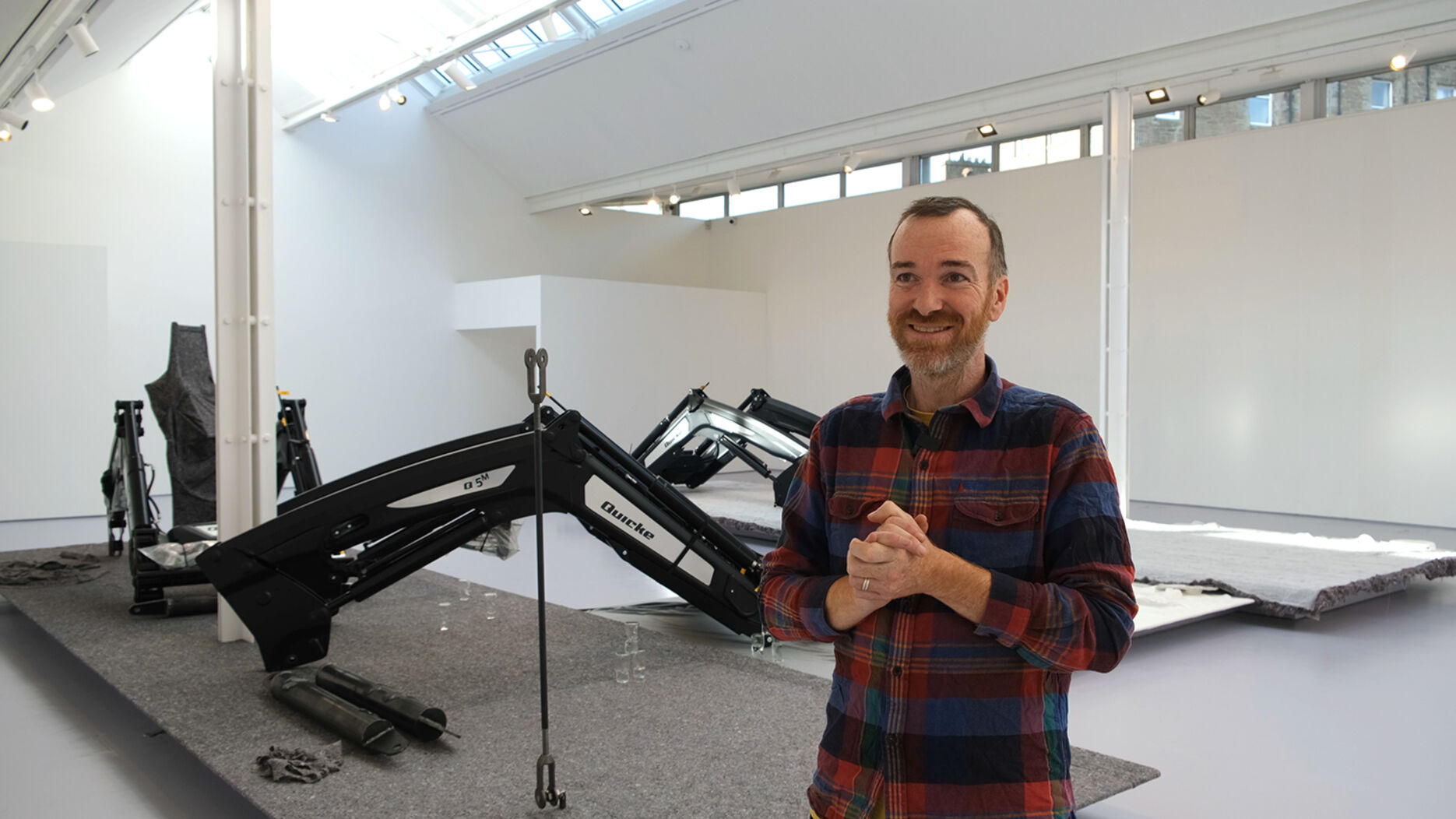 A man with a close shaven beard and a checked shirt stands in a gallery space with large farming equipment behind him