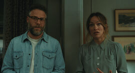 wo people stand indoors in a softly lit room, wearing casual clothing and facing forward, with framed artwork and curtains visible in the background.