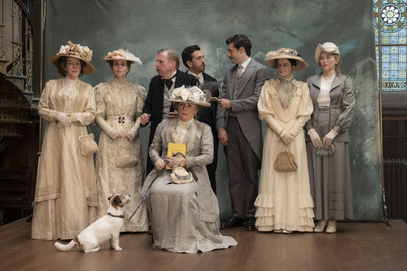 A group of people in Edwardian-style clothing pose for a formal portrait on a stage-like set, with a small dog sitting at the front and a painted backdrop and stained-glass window behind them.
