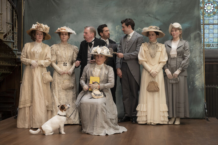 A group of people in Edwardian-style clothing pose for a formal portrait on a stage-like set, with a small dog sitting at the front and a painted backdrop and stained-glass window behind them.