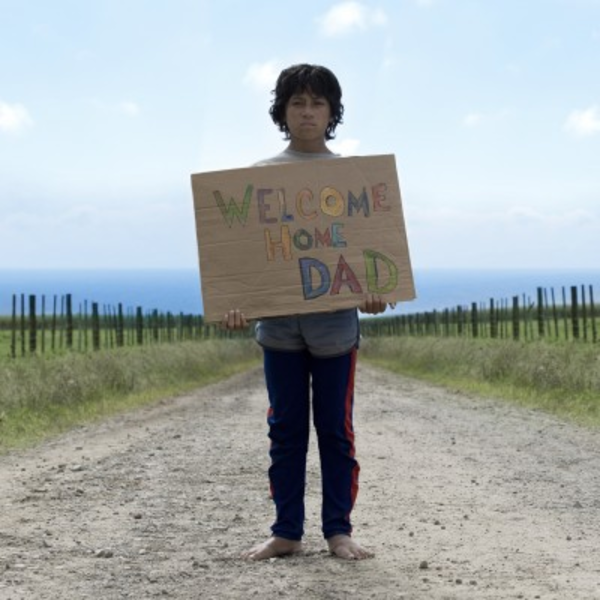 A boy stands on a dusty road holding a handmade sign reading 'Welcome Home Dad'