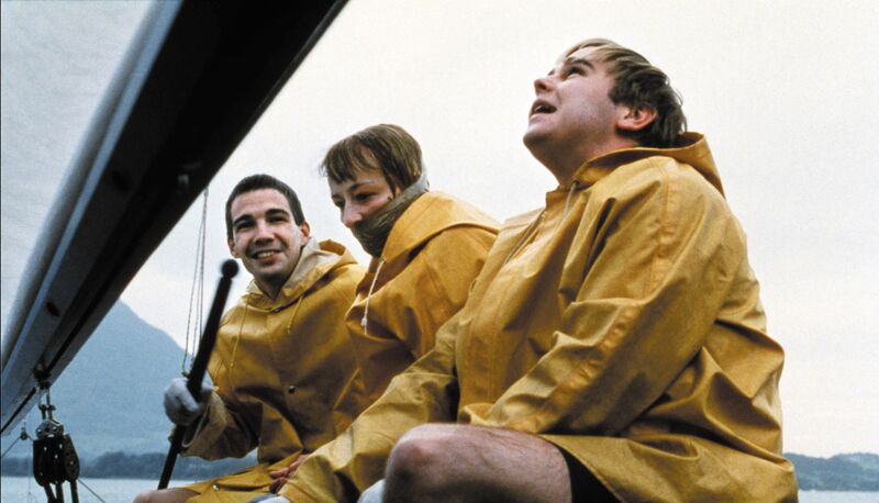 A man wearing a yellow raincoat sits on a boat with two other people wearing yellow raincoats, who look distressed