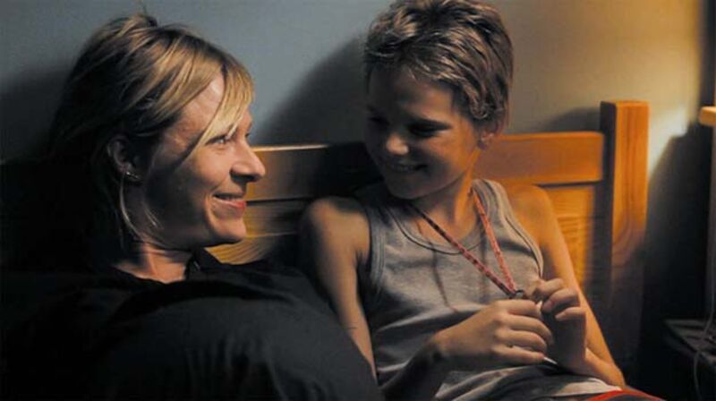 A child with short hair speaks to a woman while sitting on a bed.
