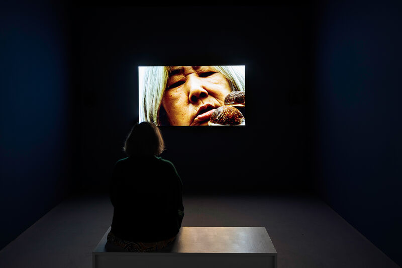 This image shows a dark gallery with a video projection. In front of the screen, a person sits on a low bench watching the film with their back to us. In the film, the face of an older woman is shown close up. The person is looking at their index finger, but we can only see part of their hand to the left side of the screen, 