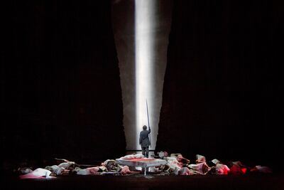 An opera singer stands with a sword, surrounded by fallen bodies, on a dramatically lit stage.