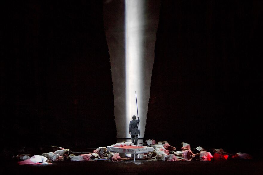 An opera singer stands with a sword, surrounded by fallen bodies, on a dramatically lit stage.