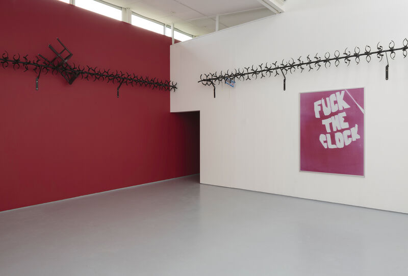 Installation photograph taken facing the intersection of two walls, at which point there is a dark doorway. The wall on the right is white and a big artwork with the words in white text 'Fuck the clock'. The wall on the left is painted red. About two thirds up the wall, installed horizontally there is black barbed metalwork.