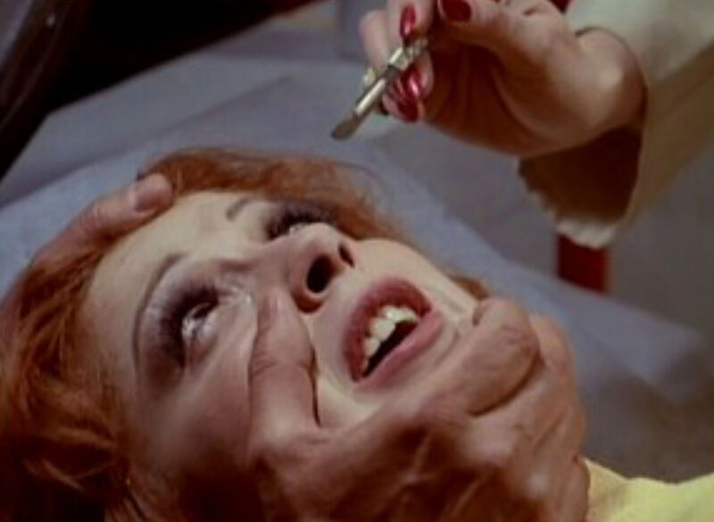 Woman lying done as someone holds her head and another woman approaches with a scalpel