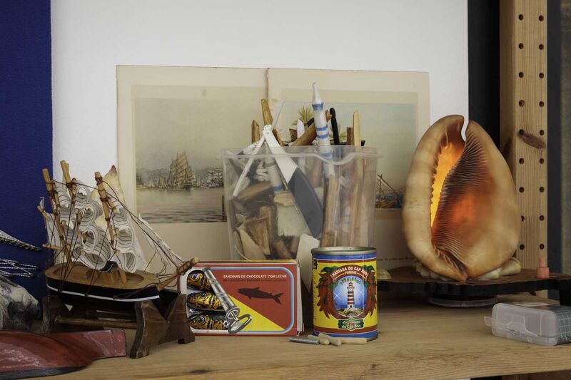 Items on a shelf, including a model boat, sardine tin, a lamp made of a large shell and a print of ships at sea.