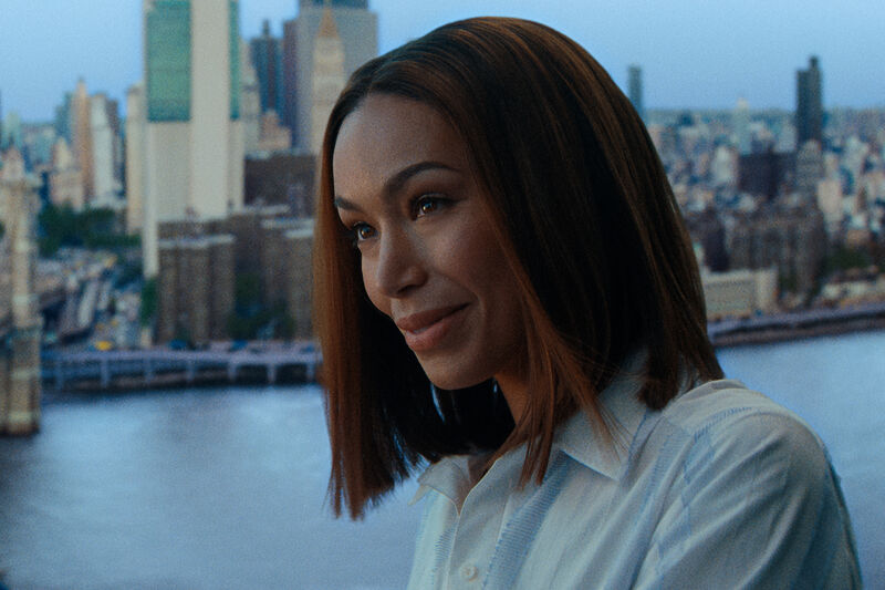 A woman with bobbed dark hair smiles by a window overlooking a cityscape.