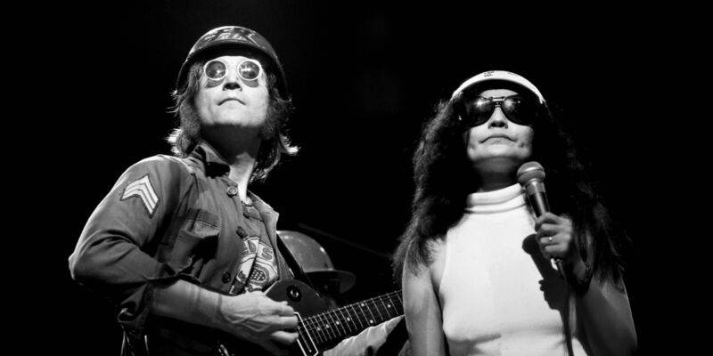 John Lennon and Yoko Ono perform on stage. John is holding a guitar and they are both wearing helmets