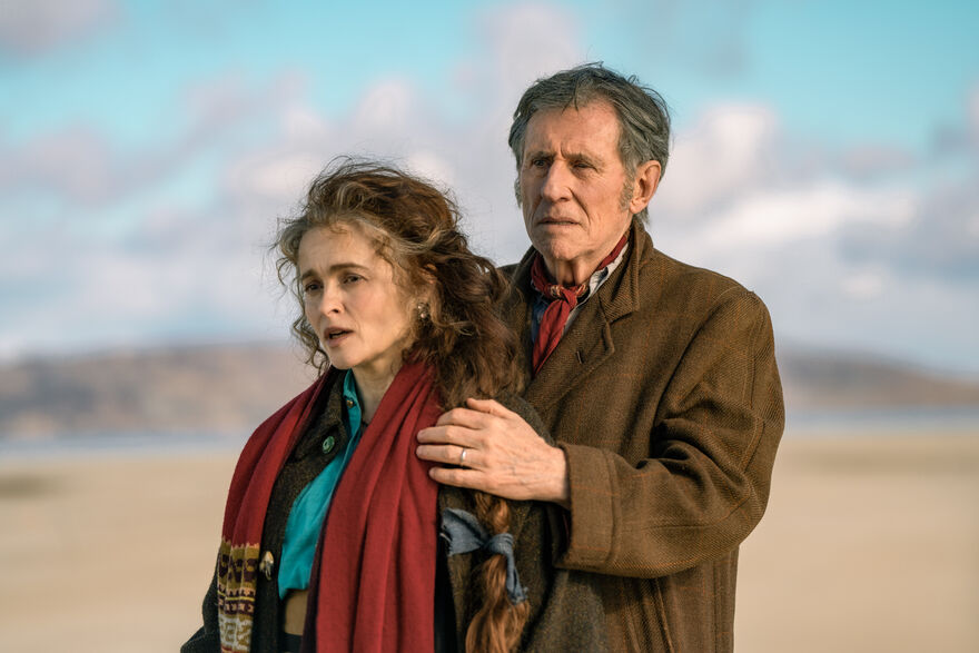 A middle-aged man and woman wearing coats and scarves stand on a beach together.