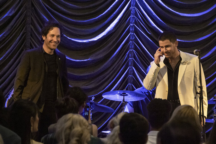 Two men stand on stage in suits, close to a drum kit.