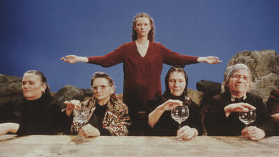 A young woman stands with her arms outstretched, behind four elderly ladies dressed as babushkas touching wine glasses.