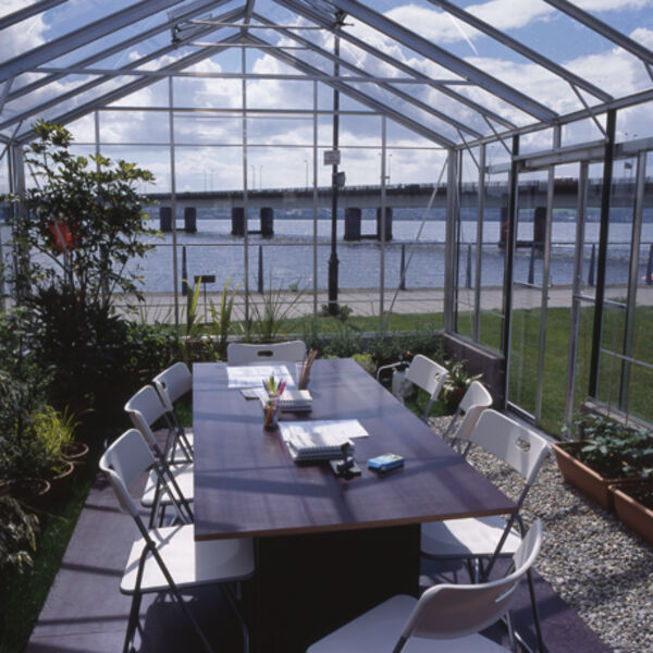 Selected works from Our Surroundings. In one photo, a glass conservatory sits on a square of grass outside in Dundee. In another, a blue image can be seen on a projector in DCA galleries. In another, an image taken from within the glass conservatory shows tables and chairs, and a view of the Tay Bridge from outside.