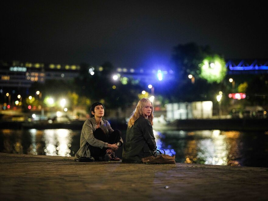 Two women sit by a river in a city at night