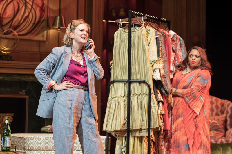 A lady on a mobile phone stands next to a rack of clothes another lady peers round the side 