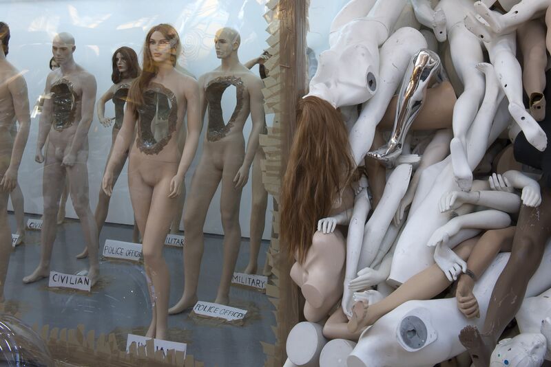 Naked mannequins are displayed with signs beneath them that say either 'police officer', 'military' or 'civillian'. The mannequins are hollowed out in the middle. Next to the standing mannequins, there is a pile of deconstructed mannequins lying in a heap.