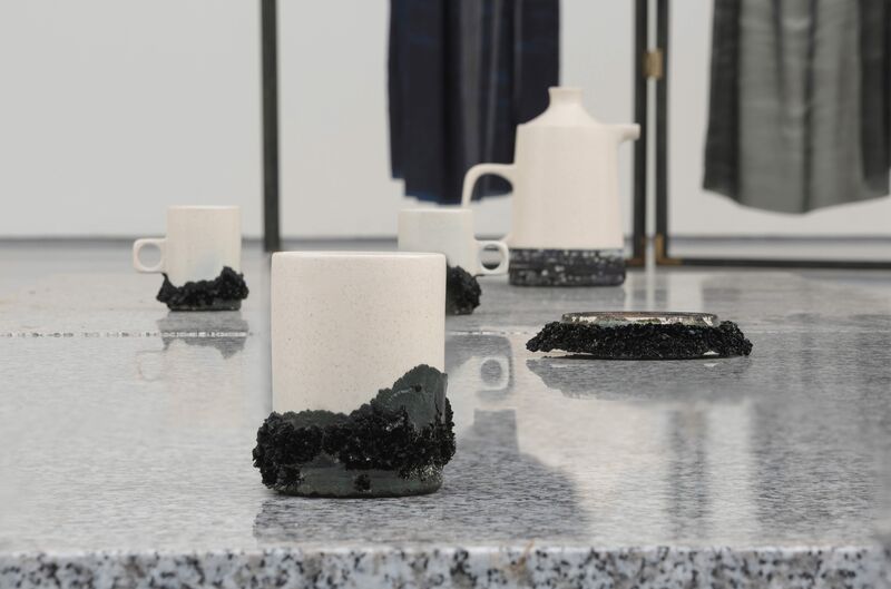 A close up from 'Pieces of You Are Here' shows white ceramic mugs and jugs with black bases.