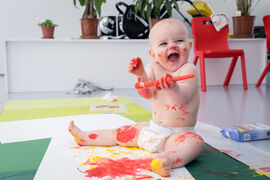 Baby smiling while holding a paint brush and covered in paint
