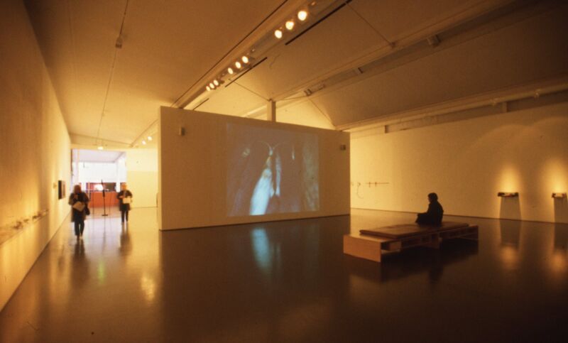 DCA galleries during Will Maclean's exhibition. The lighting in the galleries is low, and someone is sitting on the bench watching a film on a projector.