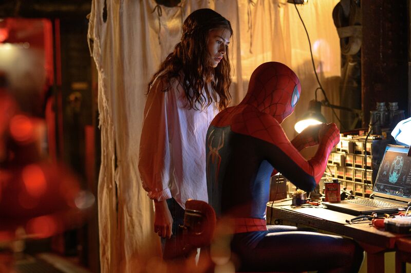 Spider-Man types on a laptop at a desk while a woman in a pink shirt stands behind him, observing his work.
