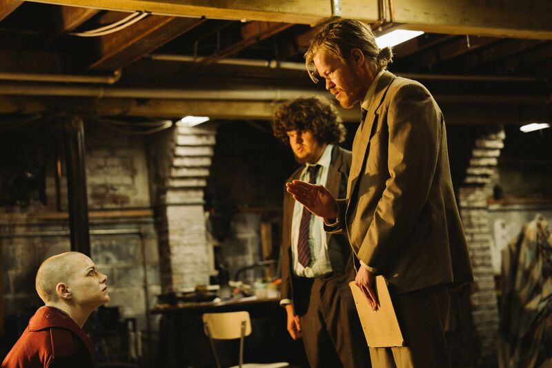Two unkempt men wearing suits talk to a woman with a shaved head in a basement