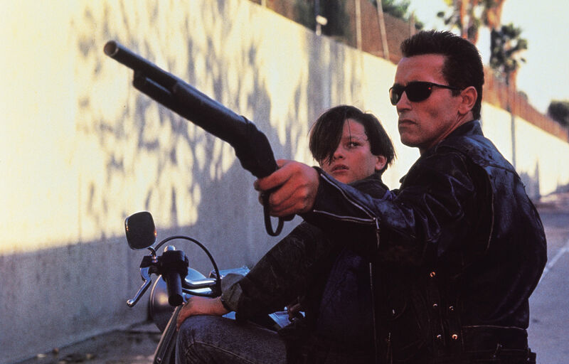 A teenage boy and a man on a motorcycle. The man is aiming a shotgun offscreen