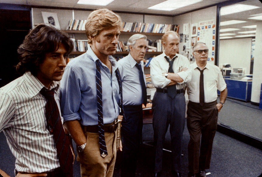 Five people in office attire stand together in a newsroom or editorial office, surrounded by bookshelves, desks and wall displays, with several wearing loosened ties and rolled-up sleeves.