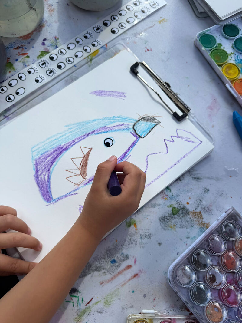 A child's had drawing with purple crayon onto paper that is on a clipboard. The table is messy with paint and eye stickers to one side 