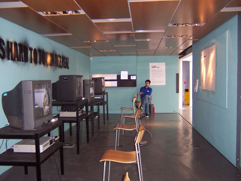 Chairs face televisions that each playing different videos. The walls are blue with the words 'It Is Hard to Touch the Real' spray painted on them.