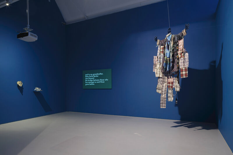 Photograph showing an installation in gallery one. Two small objects mounted on the wall are seen to the right. A screen is in the back corner showing text. A costume hangs away from the wall to the left, with fragments of fabric such as tartan. The walls are bright blue and the floor is grey, the objects are spot lit and a projector can be seen hanging from the ceiling. 