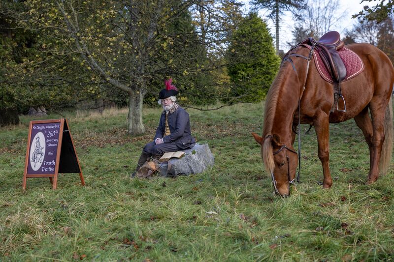 A man in historical costumes sits next to a horse.